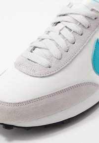 Bílé tenisky s šedými semišovými akcenty, světlými modrými detaily a texturovanými tkaničkami. Mají hladký textilní svršek a gumovou podrážku.