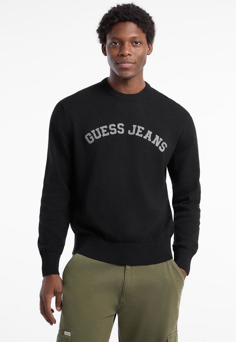 Černý pletený svetr s vyšitým nápisem "GUESS JEANS" v šedé barvě napříč hrudí. Má kulatý výstřih a žebrované manžety a lem.