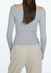 Kobieta w dopasowanym, odsłoniętym ramionom, długim szarym swetrze z prążkowanej tkaniny oraz beżowych spodniach z wysokim stanem, widziana z tyłu.