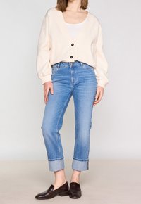 Ljus krämfärgad överdimensionerad cardigan med tre svarta knappar, parat med ljusblå raka jeans med uppvikta ben och mörkbruna loafers.