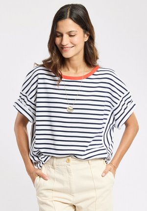 Femme aux cheveux bruns mi-longs portant une chemise rayée blanche et marine avec un col rouge et un pantalon beige, mains dans les poches, souriante en regardant vers le bas.