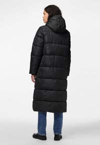 Casaco acolchoado preto longo com capuz, apresentando um design matelassado e costuras verticais. Combinado com jeans azul escuro e calçado preto.