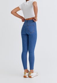 Jean skinny bleu en denim taille haute avec poches arrière, porté par un mannequin vêtu d'un top court blanc et de baskets.