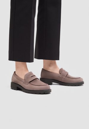Persona che indossa mocassini in camoscio colore taupe con suole nere spesse e pantaloni neri cropped, in piedi su un pavimento grigio chiaro.