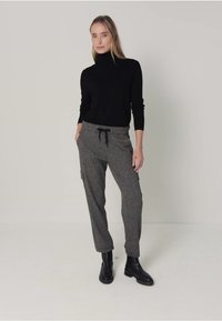 Vrouw in een zwarte coltrui, grijze broek met een trekkoord en cargozakken, en zwarte enkellaarsjes, staande tegen een effen achtergrond.