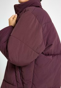 Giacca a vento bordeaux con colletto alto, texture trapuntata e zip frontale. Presenta un design aderente e una finitura morbida e opaca.