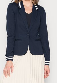 Granatowa marynarka z białymi paskami na kołnierzu i mankietach, z pojedynczym zapięciem na guzik i dwoma przednimi kieszeniami. Miękki materiał.