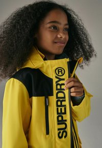 Geltona nepralaidi striukė su juodais akcentais, turinti išryškintą "SUPERDRY" logotipą, užtraukiamą kišenę ir tekstūruotą audinį.