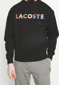 Czarna bluza z haftowanym wielokolorowym napisem "LACOSTE", ściągaczami na mankietach i dole. Subtelne logo na piersi. Miękki materiał.