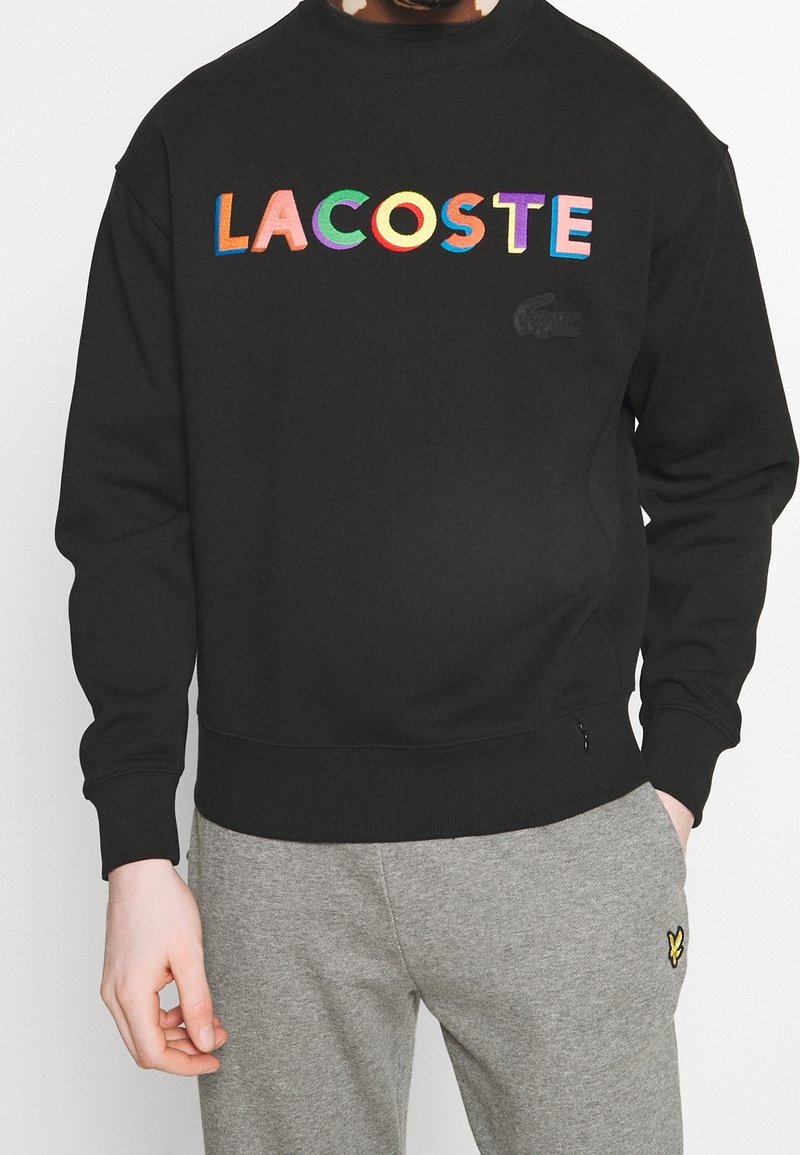 Czarna bluza z haftowanym wielokolorowym napisem "LACOSTE", ściągaczami na mankietach i dole. Subtelne logo na piersi. Miękki materiał.