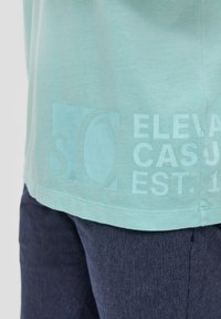 Lichtblauwe katoenen t-shirt met een ronde halslijn, voorzien van een grote grafiek in lichterblauwe tekst en vormen aan de linkerkant.