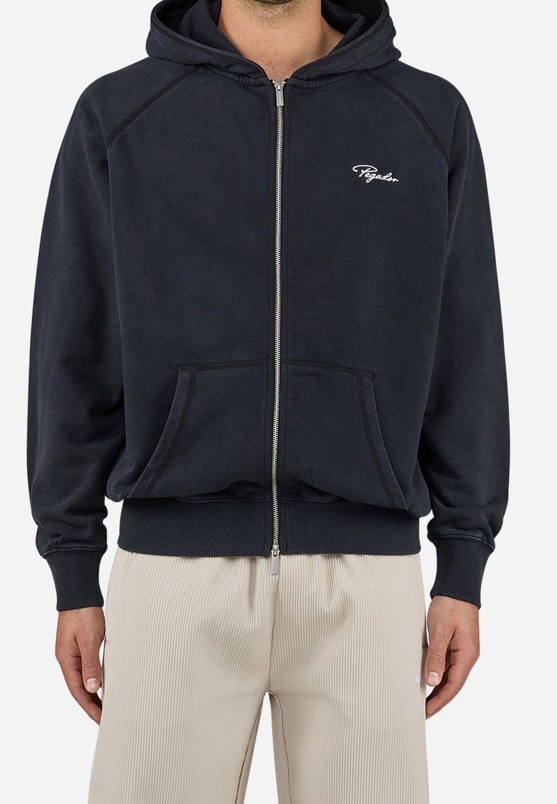 Homme portant un sweat à capuche noir zippé avec des poches avant et un pantalon côtelé beige, debout devant un fond uni.