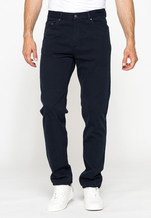 Uomo con jeans blu scuro e sneakers bianche in piedi davanti a uno sfondo neutro con le braccia rilassate ai lati.