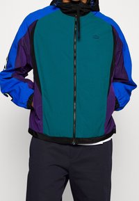 Colorful windbreaker featuring teal, blue, and purple panels. Includes a black zip front, adjustable hood, and elastic cuffs.
