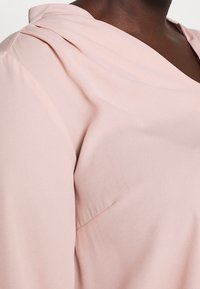 Jasnoróżowa bluzka z dekoltem w kształcie drapowania i plisowanym wzorem na ramionach. Wykonana z miękkiego materiału o gładkiej fakturze i delikatnym połysku.