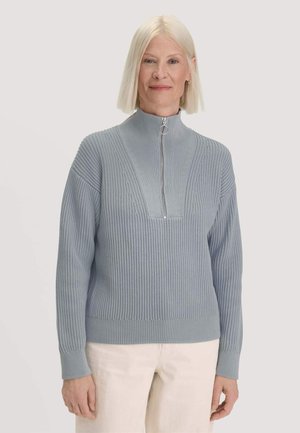 Femme aux cheveux blancs et raides portant un pull côtelé bleu clair avec une fermeture éclair à l’avant et un pantalon couleur crème.