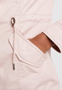 Chaqueta de color rosa claro con un bolsillo visible y una tapa diagonal, hecha de tela texturizada, mostrada con una mano descansando dentro del bolsillo.