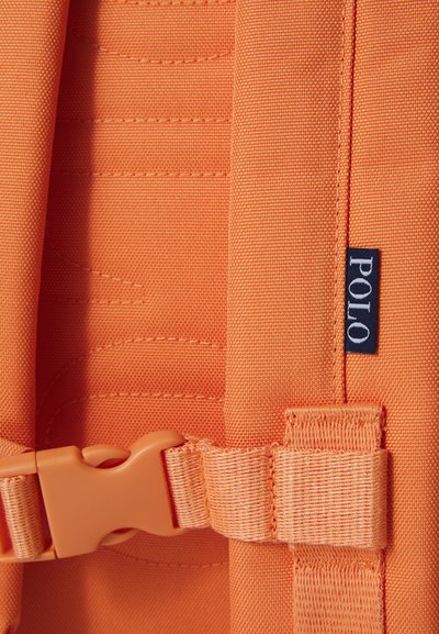 Dettaglio di uno zaino in tessuto arancione con fibbia e cuciture accentuate; piccola etichetta "POLO" in blu scuro sul lato.
