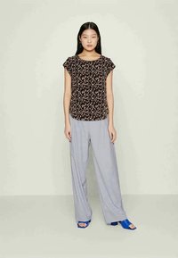 Blusa preta com padrão floral, mangas curtas, combinada com calças largas cinza claro e sandálias azuis, de pé contra um fundo neutro.