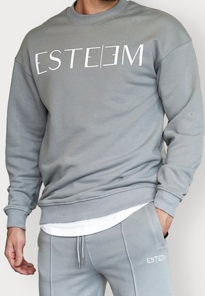 Man draagt een grijze sweatbroek en een grijze trui met het woord "ESTEEM" in het wit gedrukt, handen ontspannen, één in de zak.