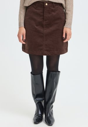 Femme portant une jupe en velours côtelé marron, des collants noirs, des bottes noires montantes et un pull beige, debout devant un fond uni.