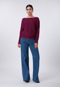 Μπορντό ριμπ πλεκτό πουλόβερ με φαρδιά λαιμόκοψη τύπου βαρκούλα, συνδυασμένο με φαρδιά μπλε τζιν και μαύρα μποτάκια με τακούνι.