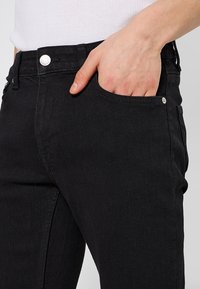 Jeans de mezclilla negra con un corte ajustado, diseño de cinco bolsillos y herrajes metálicos plateados en el bolsillo delantero y la cintura. Textura suave.