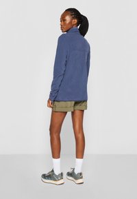 Marineblå fleecejakke med høy krage, kombinert med olivengrønne shorts. Modellen har på seg grå sneakers med oransje detaljer og hvite sokker.