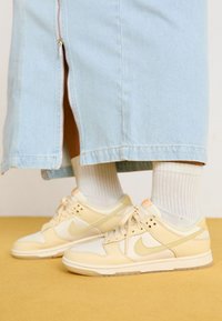 Nike Dunk-sneakers i creme og lys beige, med struktureret læder og mesh, med et minimalistisk design og en blød gummisål. Hvide sokker er synlige.