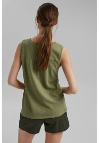 Femme aux longs cheveux bruns attachés en queue de cheval, portant un haut sans manches vert olive et un short vert foncé, debout devant un fond uni.