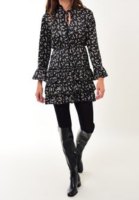 Schwarzes Blumenkleid mit langen Ärmeln und Rüschenbesatz am Saum. Hat einen gebundenen Ausschnitt und wird mit glänzenden schwarzen knielangen Stiefeln kombiniert.