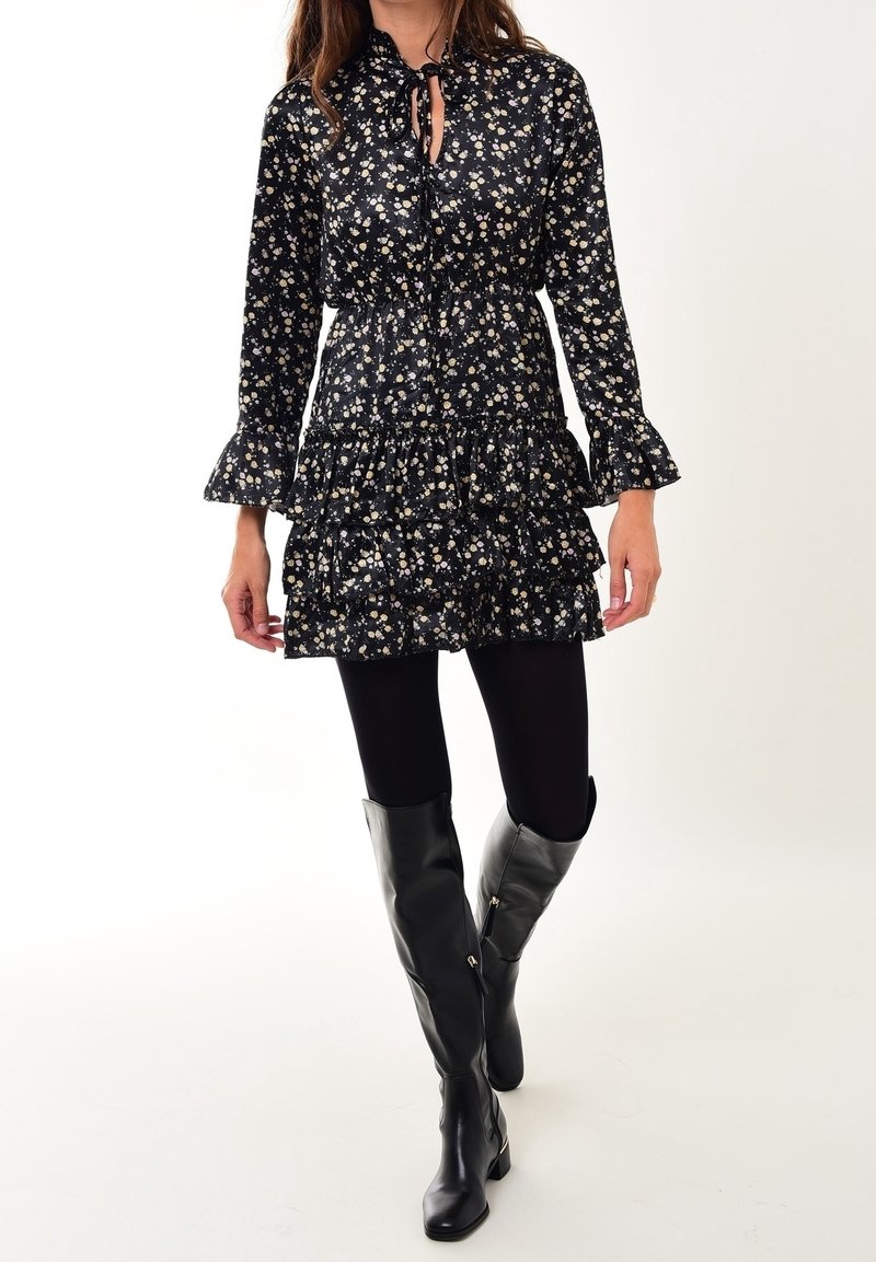 Schwarzes Blumenkleid mit langen Ärmeln und Rüschenbesatz am Saum. Hat einen gebundenen Ausschnitt und wird mit glänzenden schwarzen knielangen Stiefeln kombiniert.
