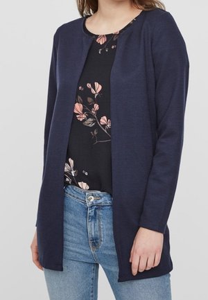 Femme portant un cardigan bleu marine ouvert sur un chemisier noir à fleurs et un jean bleu clair sur un fond uni.