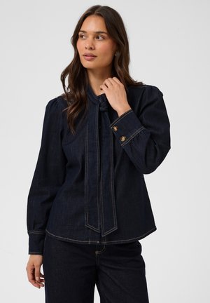 Femme aux longs cheveux foncés portant une blouse en denim foncé avec un nœud noué au col et un pantalon en denim foncé assorti sur un fond uni.