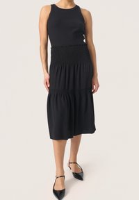 Schwarzes Kleid mit einem enganliegenden, gerippten Oberteil und geraffter Taille, dazu ein fließender, gestufter Rock. Getragen mit schwarzen Schuhen mit spitzem Zehen.