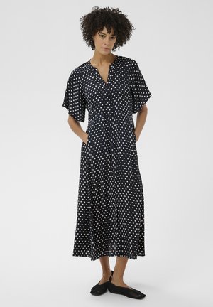 Femme aux cheveux bouclés portant une robe mi-longue noire à pois blancs, manches courtes, boutons à l'avant, et chaussures noires en maillage à enfiler.