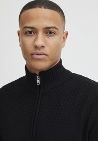 Junger Mann mit kurzen Haaren, der einen schwarzen strukturierten Reißverschluss-Pullover mit geripptem Kragen trägt und direkt in die Kamera schaut.