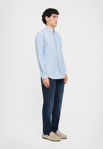 Cămașă butonată de culoare albastră deschisă, cu guler, mâneci lungi și un buzunar pe piept, purtată cu blugi slim-fit de culoare bleumarin și pantofi slip-on bej.