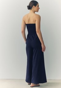 Mørkeblå ballon jumpsuit uden stropper med rynket bodice og bred ben-design. Blødt stof med subtil tekstur; har sidelommer.