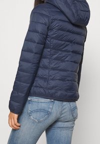 Marinblå pufferjacka med huva, som har sydda sektioner för isolering och en slät, något glänsande yta. Matchas med ljusblå jeans.