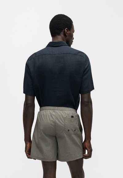 Άνδρας που φαίνεται από πίσω, φορά κοντομάνικο σκούρο πουκάμισο μέσα σε ανοιχτό γκρι σορτς με ελαστική μέση και πίσω τσέπη.