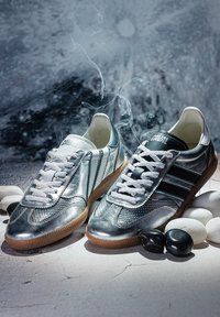 Paire de chaussures de sport argentées brillantes avec des lacets blancs et des côtés rayés sur une surface fissurée avec des pierres noires et blanches à proximité.