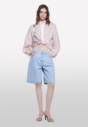 Giovane donna che indossa una camicetta rosa a righe annodata, pantaloncini di denim blu chiaro fino al ginocchio, tacchi argento e una borsa a tracolla argento.