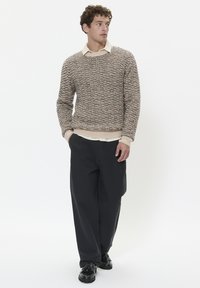 Pulover tricotat în nuanțe de bej și crem cu un model texturat, purtat peste o cămașă cu guler, asortat cu pantaloni întunecați, largi și pantofi negri.