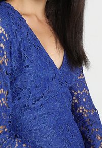 Haut en dentelle bleue avec un décolleté en V et des manches longues. Le motif complexe de la dentelle comprend des dessins floraux et circulaires avec des mailles ajourées.