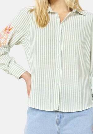 Femme portant une chemise à rayures verticales vert clair et blanc avec une broderie florale sur la manche gauche, associée à un jean bleu clair.