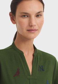 Chemise verte avec col montant, texture subtile et designs brodés ludiques incluant des animaux et des lettres de différentes couleurs.