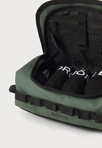 Zelena torba sa patentnim zatvaračem i teksturiranim unutrašnjim dijelom sadrži tri presavijena crna predmeta, ima standardne hardverske komponente i brendiranje na vanjskoj strani.