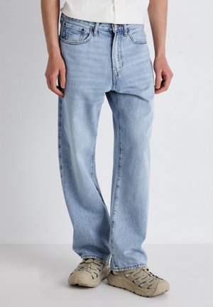Personne portant un jean droit bleu clair et des baskets beiges debout devant un fond clair uni.