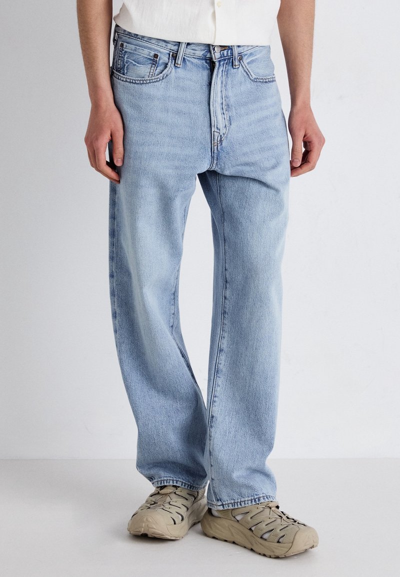 Personne portant un jean droit bleu clair et des baskets beiges debout devant un fond clair uni.